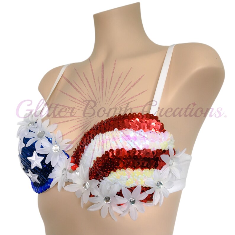 May include: A bra featuring a red, white, and blue sequined pattern resembling the American flag. The left cup is blue with white stars, while the right cup has red and white stripes. White fabric flowers with center crystals are attached.
