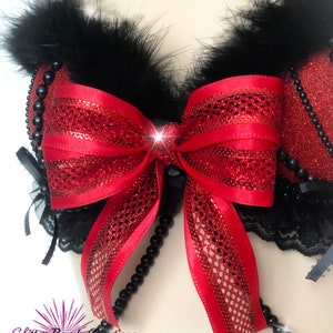 May include: A red and black lingerie top with a large red bow and black feather trim. The top is embellished with black beads and lace. The image has a sparkly effect.