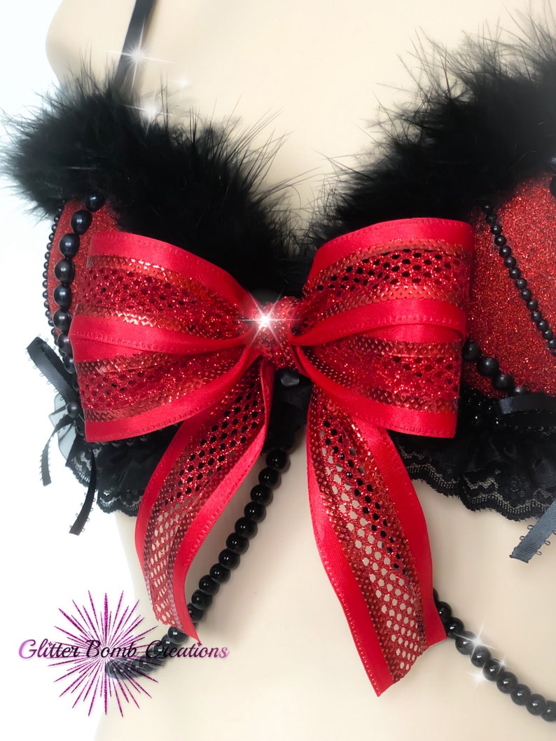 May include: A red and black lingerie top with a large red bow and black feather trim. The top is embellished with black beads and lace. The image has a sparkly effect.