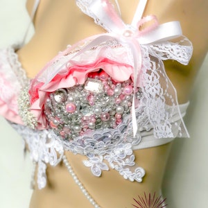 Op de afbeelding: Een witte en roze lingerie bh met kant en parelversiering. De bh heeft een wit lint dat bovenaan in een strik is gebonden. De bh is versierd met roze en witte ruches en een verscheidenheid aan parels en kralen.