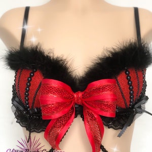 May include: Red and black embellished bra with a large red bow. The bra features black feathers, beaded accents, and black lace trim. The bra has thin black straps and a beaded chain.