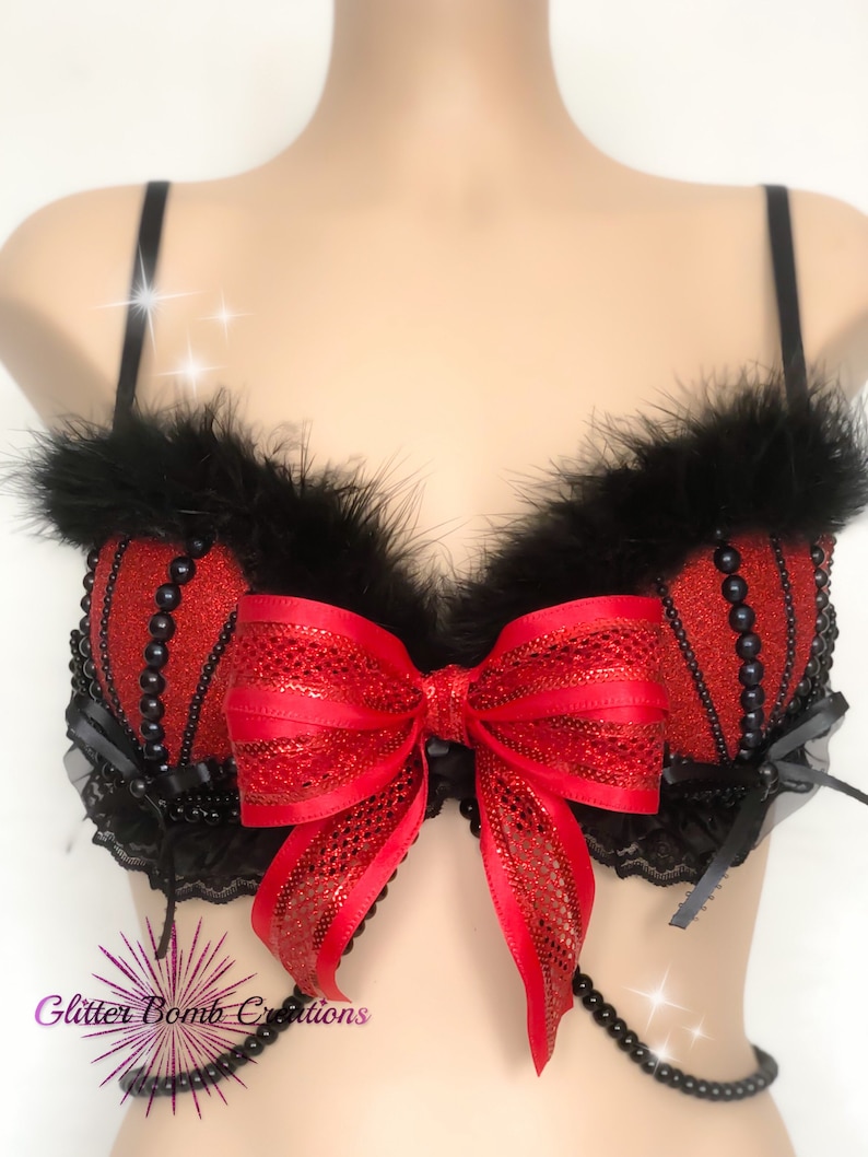 May include: Red and black embellished bra with a large red bow. The bra features black feathers, beaded accents, and black lace trim. The bra has thin black straps and a beaded chain.