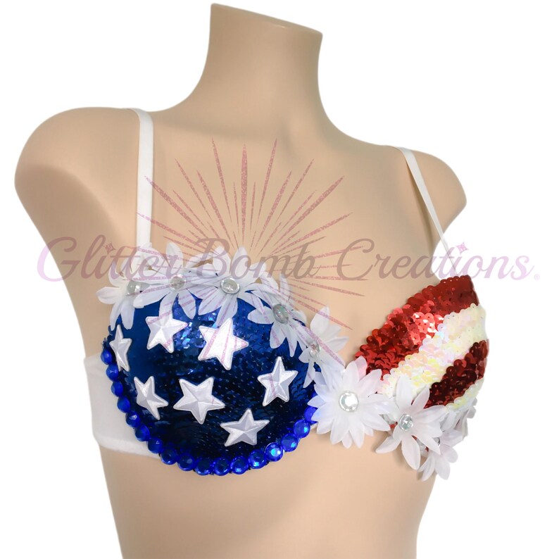 May include: A white bra featuring a blue sequined cup adorned with white stars and white fabric flowers, and a red and white striped sequined cup decorated with white fabric flowers. The text Glitter Bomb Creations is visible on the image.
