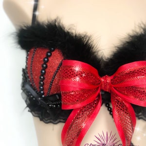 May include: Red and black lingerie set with a large red bow and black feather trim. The bra features red glitter fabric and black bead accents. The set includes a beaded chain and black lace detailing. The text "Glitter Bomb Creations" is visible.