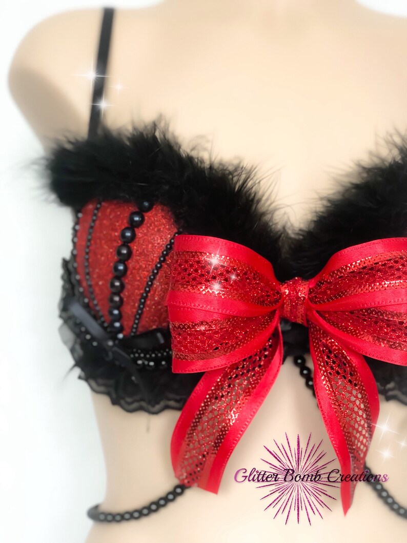 May include: Red and black lingerie set with a large red bow and black feather trim. The bra features red glitter fabric and black bead accents. The set includes a beaded chain and black lace detailing. The text "Glitter Bomb Creations" is visible.
