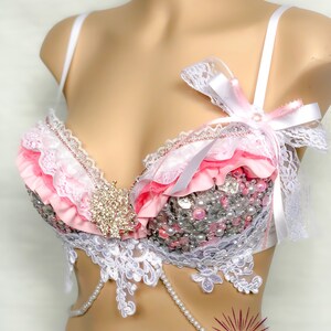 May include: A white and pink lace bra with a pearl and rhinestone embellishment. The bra has a white satin bow on the strap.