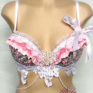 Op de afbeelding: Een roze en witte bh met kant en parelversiering. De bh heeft een groot strass-versiering in het midden. De bh is op een wit mannequin.