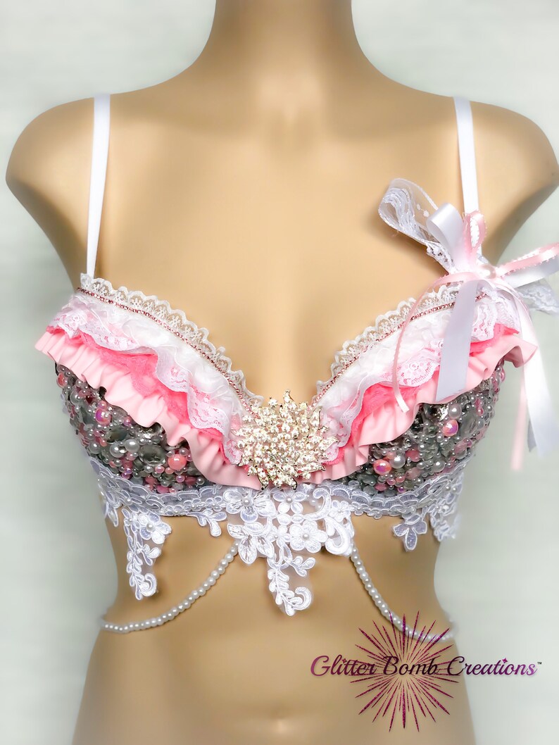 Op de afbeelding: Een roze en witte bh met kant en parelversiering. De bh heeft een groot strass-versiering in het midden. De bh is op een wit mannequin.