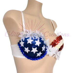 May include: A white bra featuring a blue sequined cup adorned with white stars and a red and white striped sequined cup. The garment is decorated with white fabric flowers along the top and blue gems along the bottom edge of the blue cup.