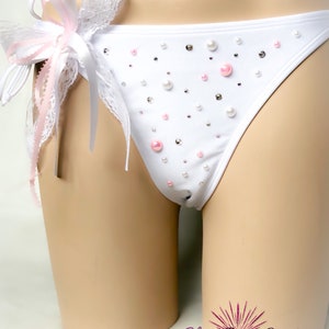 Op de afbeelding: Witte lingerie met een roze en witte kanten strik en parelversieringen. De lingerie is op een wit mannequin.
