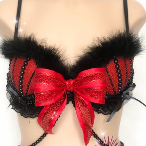 May include: A red and black embellished bra with a large red bow in the center. The bra features black feathers along the top edge and is adorned with black beads. A black beaded chain hangs from the bottom. The text "Glitter Bomb Creations" is visible.
