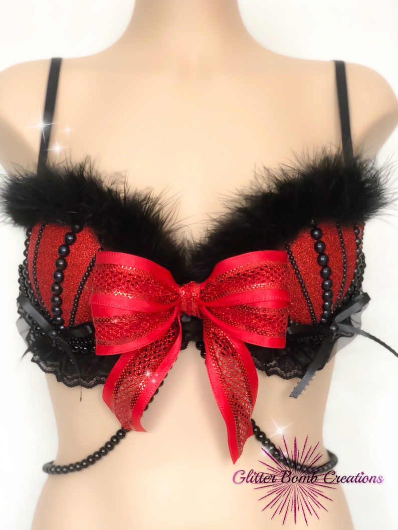 May include: A red and black embellished bra with a large red bow in the center. The bra features black feathers along the top edge and is adorned with black beads. A black beaded chain hangs from the bottom. The text "Glitter Bomb Creations" is visible.