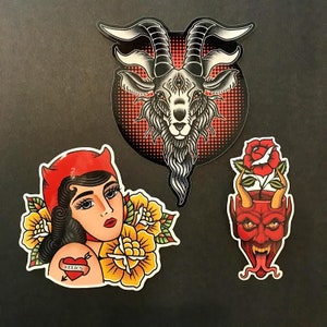 May include: Three colorful stickers with tattoo-style designs. One features a woman with a red devil horn hat and a heart tattoo that says "Relics". Another is a goat head with three eyes. The third is a red devil head with a rose.