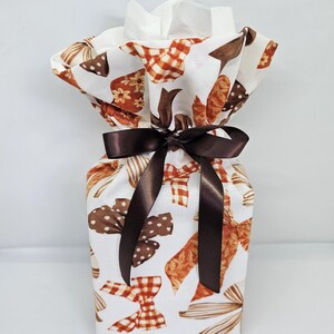 May include: A rectangular fabric tissue box cover featuring a white background with a repeating pattern of brown and orange bows in various styles, including plaid and polka dot designs, cinched at the top with a dark brown satin ribbon bow.