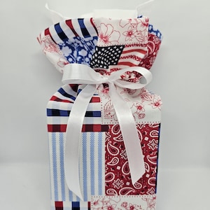 May include: A fabric gift bag with a patchwork design featuring red, white, and blue patterns. The bag is tied with a white satin ribbon. The patterns include stripes, floral prints, and a section resembling the American flag.