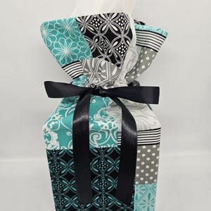 May include: A fabric gift bag with a patchwork design in teal, black, white, and gray. The bag is tied with a black satin ribbon and contains white tissue paper. The fabric features various patterns, including floral, geometric, and polka dots.
