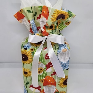 May include: A gift bag wrapped in fabric with a pattern of sunflowers, gnomes, and mushrooms on a light green background. The bag is tied with a white satin ribbon and bow. White tissue paper is visible at the top.