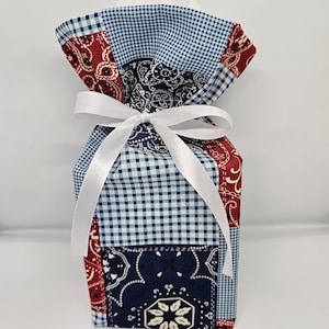 May include: A patchwork tissue box cover with a white satin ribbon. The fabric features a mix of blue gingham, paisley, and solid red and navy blue patterns. The tissue box cover is a decorative item.