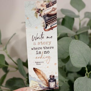 Puede incluir: Un marcapáginas blanco con una máquina de escribir vintage, una pluma y un tintero. El marcapáginas tiene el texto "Write me a story where there is no ending - Rebecca Ross".
