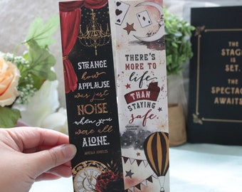 May include: Two bookmarks with quotes about life and applause. The first bookmark is black with gold accents and features a quote by Jancika Angeles: "Strange how applause was just noise when you were all alone." The second bookmark is white with red, gold, and black accents and features a quote by Stephanie Garber: "There's more to life than staying safe."