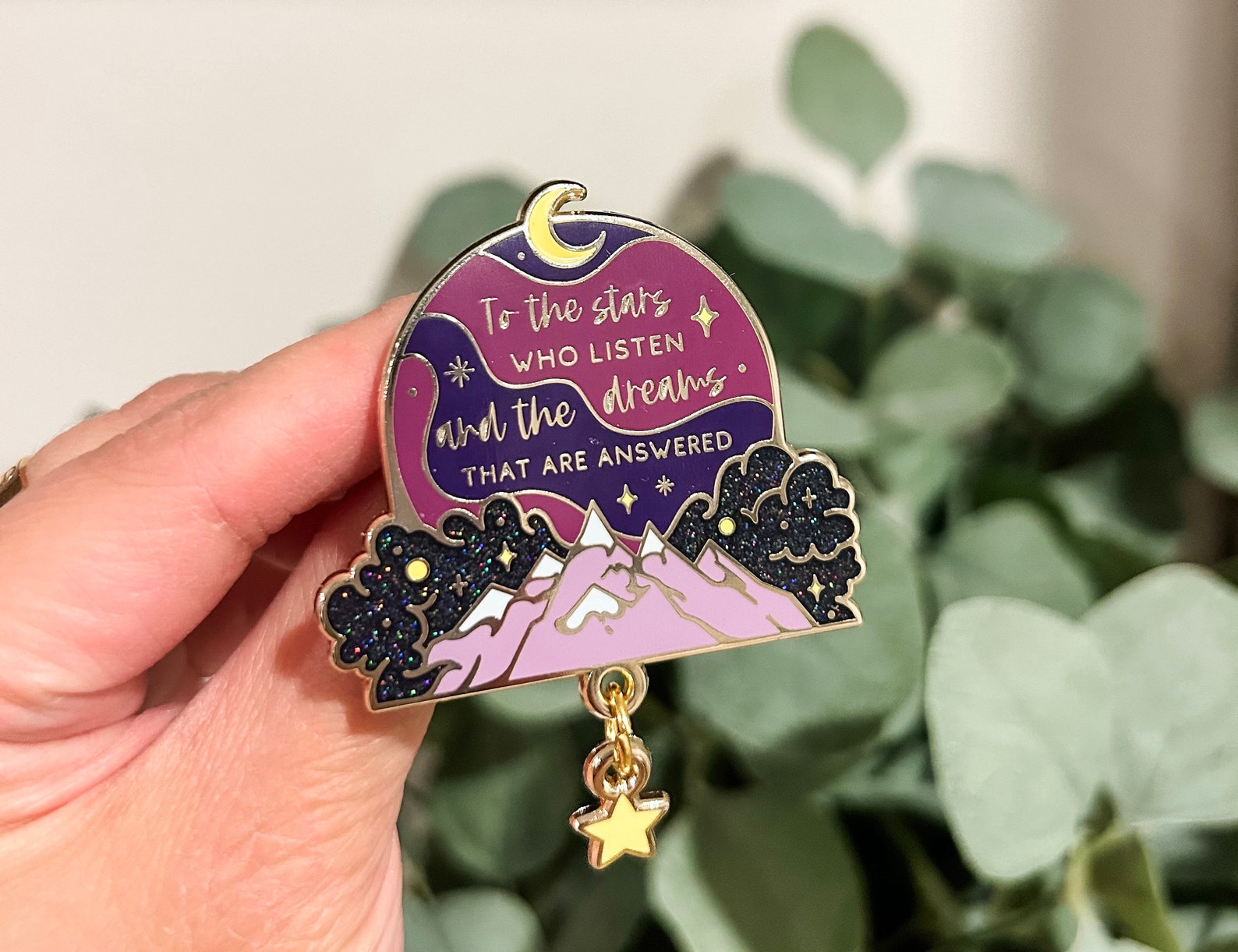 A maroon and purple pendant with the text 'To the stars who listen and the dreams that are answered' written across the circular surface. Below the text on the pendant is a shiny mountain-scape and a star pendant hanging at the bottom.