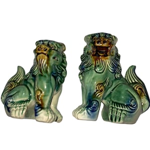 May include: Pair of ceramic guardian lion statues. The statues are in a seated position with detailed manes, tails, and paws. The lions are glazed in shades of green, blue, and brown, with intricate patterns. These decorative statues are ideal for home decor.