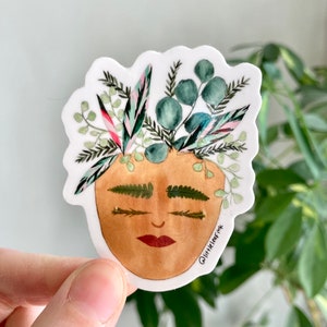 May include: A sticker featuring a watercolor illustration of a face with a plant-filled head. The face is tan with closed eyes and a red lip. The plant life includes green, pink, and blue-green leaves. The sticker has a white border and the text "@littleleafink".