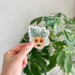 May include: A sticker featuring a watercolor illustration of a face with floral details. The face has green eyeshadow and yellow flower petals for eyes. The face is surrounded by green leaves and flowers.