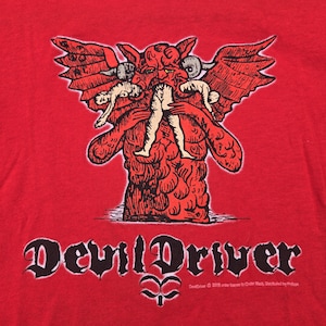 Puede incluir: Camiseta roja con una ilustración detallada de una figura roja parecida a un demonio con alas, cuernos y sosteniendo dos figuras. El texto "DevilDriver" está impreso en una fuente gótica. La camiseta es de material suave.