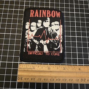 Puede incluir: Parche negro con la palabra "RAINBOW" en rojo sobre un gráfico de personas y una línea roja de latidos. El texto "DIFFICULT TO CURE" está debajo. El parche mide aproximadamente 7,6 cm de ancho.