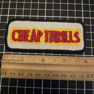 Puede incluir: Parche bordado con las palabras "CHEAP THRILLS" en letras rojas con llamas amarillas. El parche es rectangular con un borde negro y un fondo gris claro. Una regla de madera está debajo.