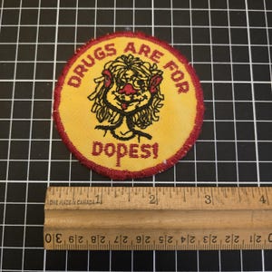 May include: A circular yellow patch with red edging, displaying a cartoon figure and the words "DRUGS ARE FOR DOPES!". The design is in black and red.