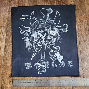 Op de afbeelding: Zwart-witte geborduurde patch met een schedel die een bandana draagt en een pijp vasthoudt. De tekst "MODEL METALLICA" en "ZORLAC" is zichtbaar op de patch.