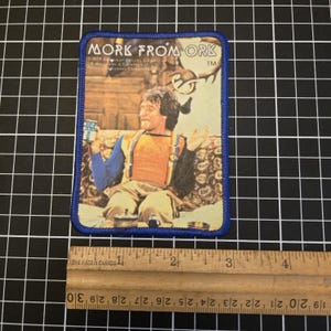May include: A rectangular patch with a blue border featuring an image of Mork from the TV show Mork & Mindy. The image shows Mork in an orange vest and blue sleeves. The text "MORK FROM ORK" is at the top.