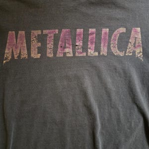 Peut inclure: T-shirt gris foncé avec le nom du groupe "METALLICA" imprimé en lettres violettes et beiges délavées. L'impression est vieillie, donnant au t-shirt un aspect vintage.