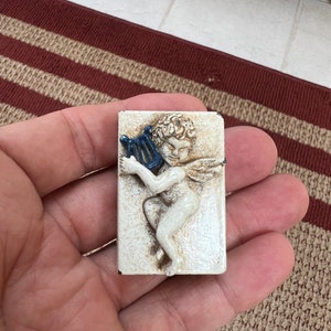 May include: A white ceramic tile with a raised relief sculpture of a cherub playing a blue lyre. The cherub is depicted with wings and a flowing hairstyle.