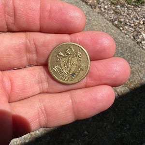 May include: A round, antique brass button with an embossed design. The button features a central figure within a shield, surrounded by text. The button is held in a hand, showcasing its size and detail.