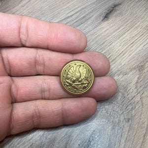 May include: A gold-colored button with an eagle design. The eagle is perched on a shield with stars and stripes. The button is about 1 inch in diameter.