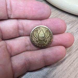 May include: A vintage brass button with an embossed eagle and shield design. The button is round and has a detailed relief of the American eagle with outstretched wings, a shield, and decorative elements. The button is held in a hand.