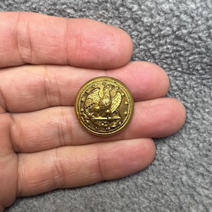 May include: A gold-colored metal button with an eagle design. The eagle is holding a bundle of arrows in its talons and has a crown on its head. The button has a raised rim and a smooth surface.