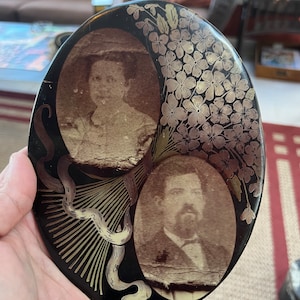 May include: A black circular trinket box with gold accents. The box features two oval portraits of a man and a woman. The portraits are surrounded by a floral design with gold leaves and flowers.