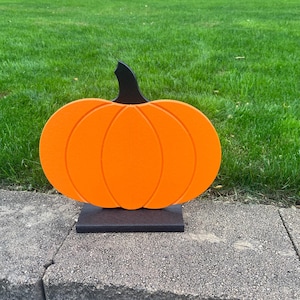 May include: A flat, orange pumpkin cutout with a black stem, featuring carved vertical lines to define the segments. The pumpkin is securely attached to a rectangular black base, providing a stable foundation for this decorative object.