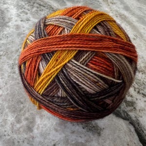 May include: A spherical ball of yarn featuring a variegated color palette of deep brown, charcoal gray, warm orange, and golden yellow. The strands are tightly wound in a crisscross pattern, showcasing a multi-tonal texture throughout the entire ball.