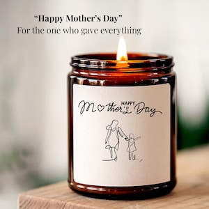 Puede incluir: Una vela encendida en un frasco de vidrio marrón con una etiqueta blanca. La etiqueta presenta un dibujo lineal de una madre y un niño, con las palabras "Happy Mother's Day" y "For the one who gave everything."