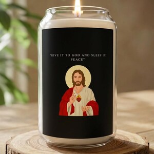Puede incluir: Una vela encendida en un tarro de vidrio transparente con una etiqueta negra. La etiqueta muestra una imagen de Jesús y el texto "GIVE IT TO GOD AND SLEEP IN PEACE." La vela está sobre una rodaja de madera.