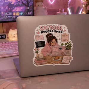 Puede incluir: Un portátil con una pegatina "SOFTGIRL PROGRAMMER" está sobre un escritorio con periféricos rosas. También se ven una vela encendida, una bebida rosa y un monitor de ordenador que muestra un paisaje urbano. La habitación está decorada en tonos rosas.
