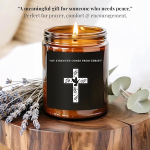 Puede incluir: Una vela encendida en un frasco de vidrio marrón con una etiqueta negra con un diseño de cruz blanca. El texto en la etiqueta dice "MY STRENGTH COMES FROM CHRIST". La imagen también incluye el texto "Un regalo significativo para alguien que necesita paz."