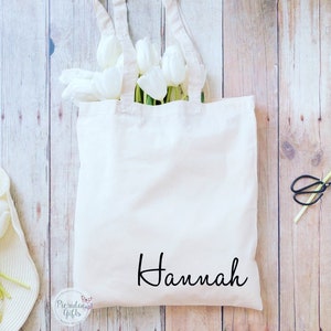 May include: White canvas tote bag with black handles and the name "Hannah" printed in black cursive script. The bag is filled with white tulips.