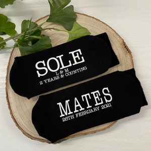 May include: A pair of black socks with white text. One sock says "SOLE L & M 2 YEARS & COUNTING" and the other sock says "MATES 26TH FEBRUARY 2021".