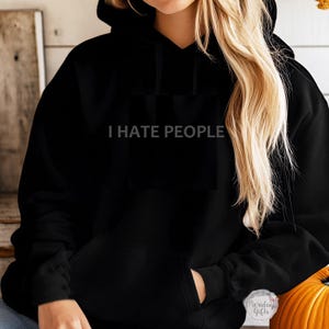 May include: Black hooded sweatshirt featuring the white text "I HATE PEOPLE". The sweatshirt has a front pocket and a hood. The model is wearing the sweatshirt.